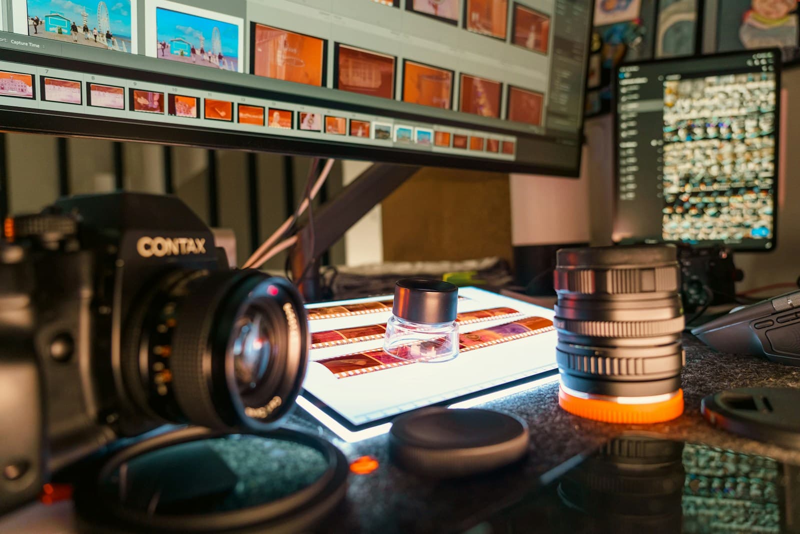 A film photography desk in a dim room: a Contax camera and lenses beside a glowing light table holding film strips and a loupe, with scans on a monitor behind