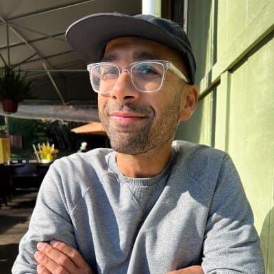 Lokesh Dhakar smiling in the sun, wearing a dark cap, clear glasses and a grey sweatshirt, arms crossed, in front of a green wooden wall